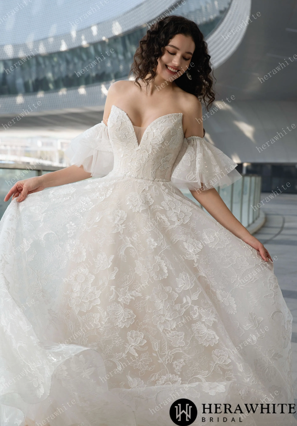 V-Neck allover lace wedding dress featuring a flowing skirt and elegant off-shoulder sleeves against a modern backdrop.