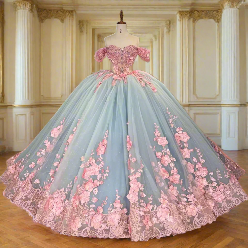A stunning blue and pink Quinceanera dress with a sheer lace bodice, 3D floral decorations, a sweetheart neckline, and a full ballroom skirt with an oversized back bow.