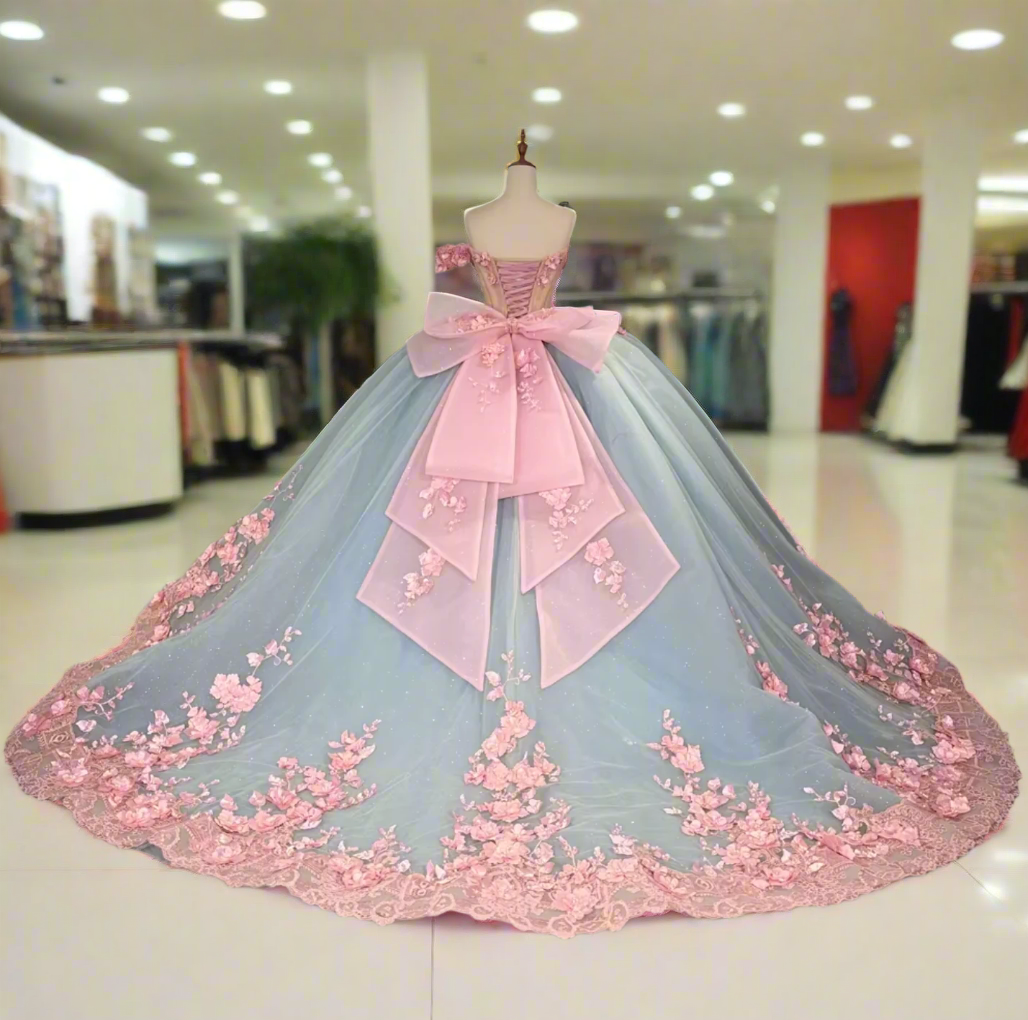 A stunning blue and pink Quinceanera dress with a sheer lace bodice, 3D floral decorations, a sweetheart neckline, and a full ballroom skirt with an oversized back bow.