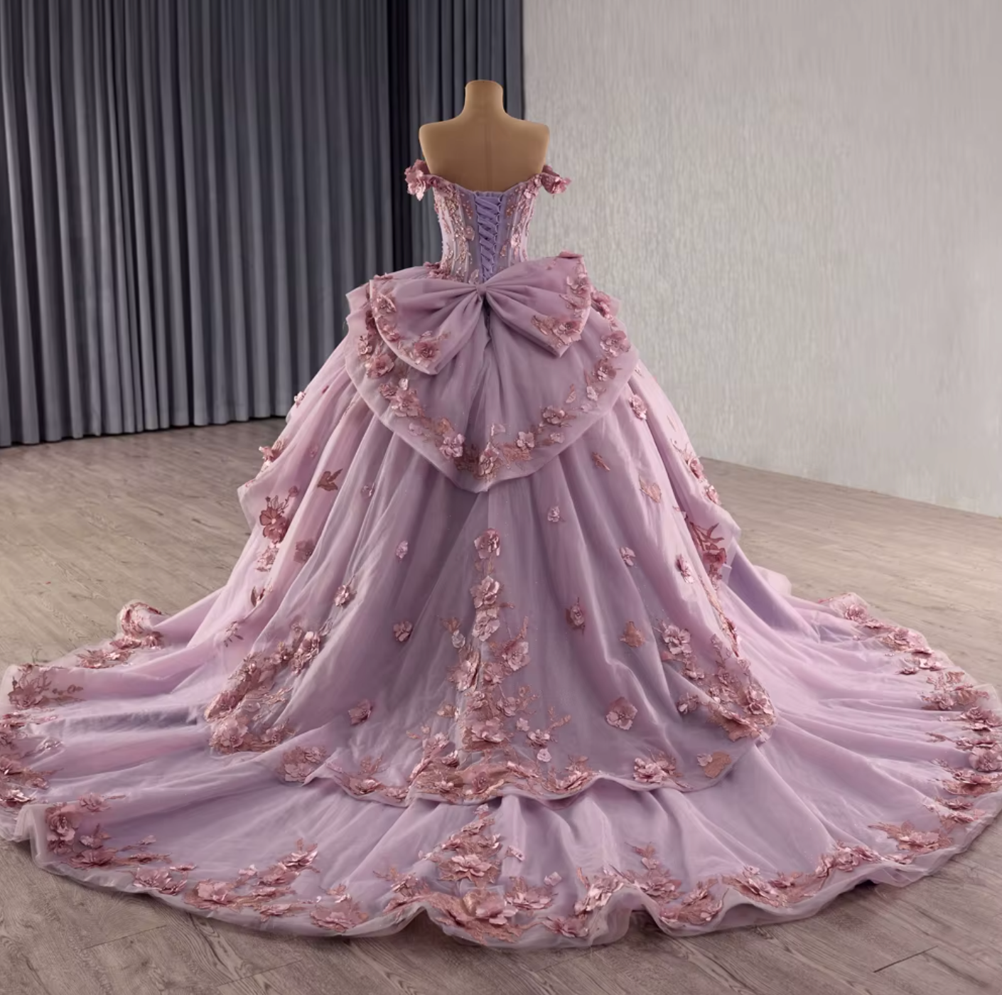 Back view of a lilac purple off shoulder quinceanera dress with lace, ruffles, and floral embellishments.